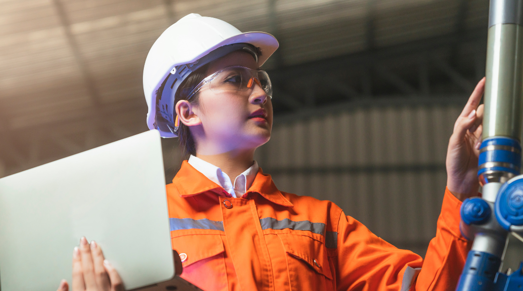 Woman with hardhat and safety glasses with laptop in one hand is checking manufacturing equipment to make sure it complies with lean manufacturing techniques
