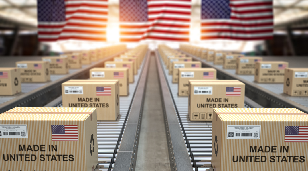 The Importance of Manufacturing in America blog feature image is boxes on conveyer belt coming towards viewer that say "Made in United States" as three American flags fly overhead.