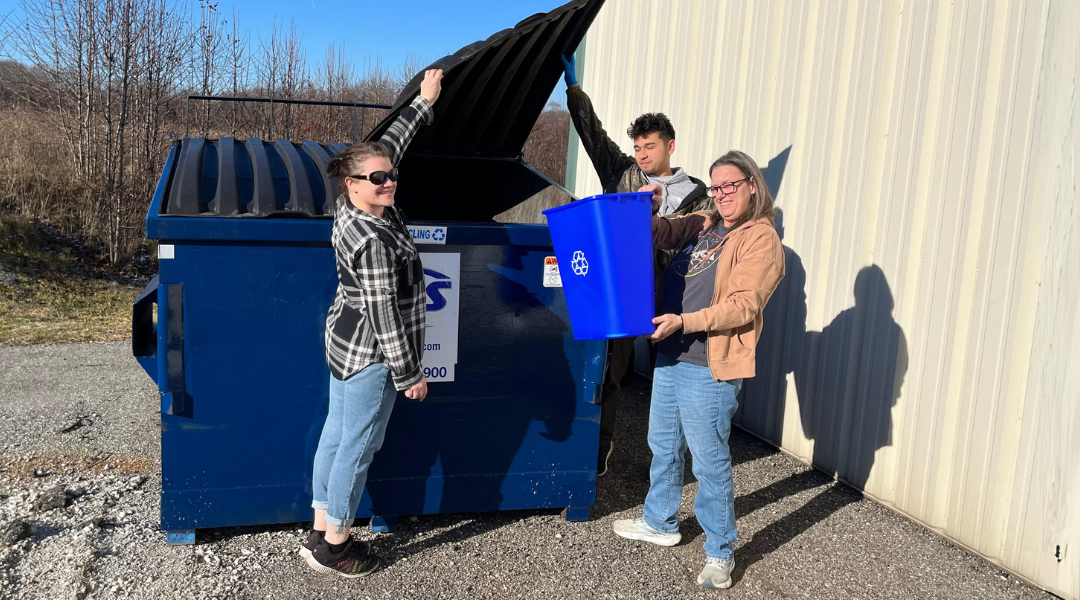 Three Winndeavor team members practice sustainable manufacturing by placing recyclable materials inside a recycle dumpster.