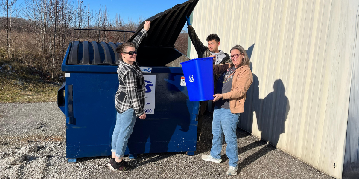 Three Winndeavor team members practice sustainable manufacturing by placing recyclable materials inside a recycle dumpster.