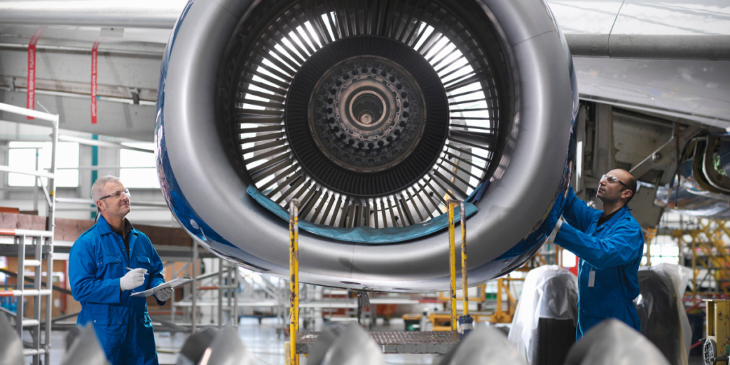 Two men stand on opposite sides of airplane turbine inspecting it.