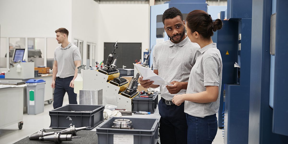 Engineer And Apprentice Meet On Floor Of Precision Machining Shop