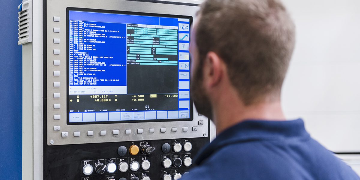 Technician monitoring a CNC machine control panel with a blue screen showing programming codes and settings.